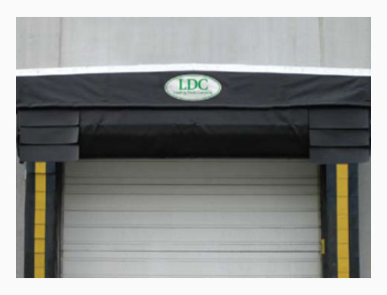 A loading dock door with a black cover and yellow safety strips on both sides, featuring loading dock seals Windsor. An oval sign above the door reads LDC against a gray concrete wall.